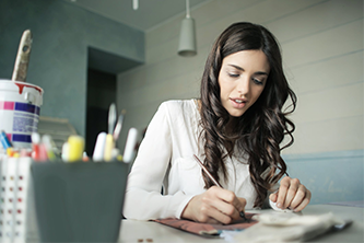 a woman with long, wavy hair sitting at a desk, writing or sketching on paper.